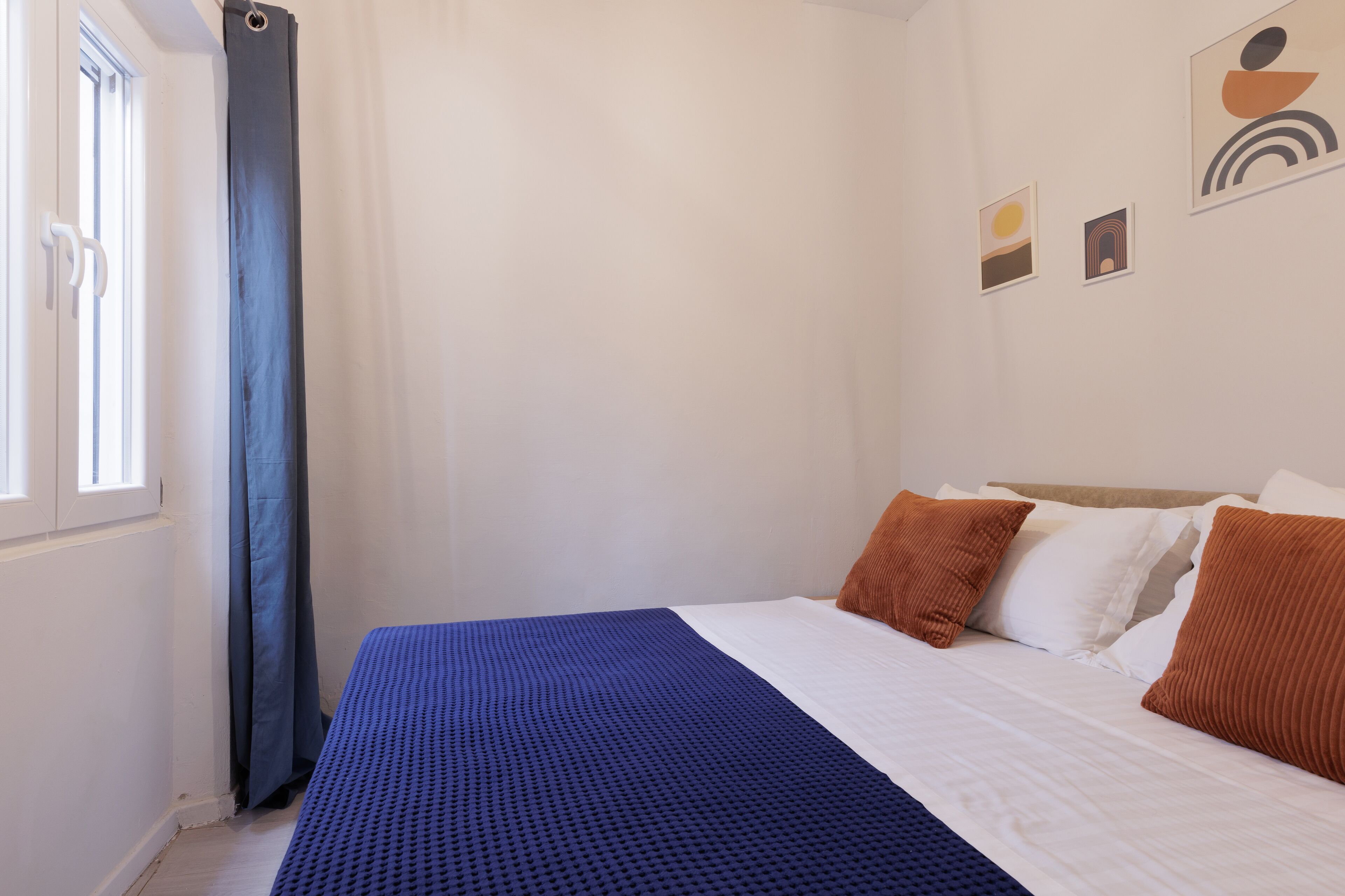 A bedroom with a bed, white walls, a window with curtains, and three small wall art pieces.