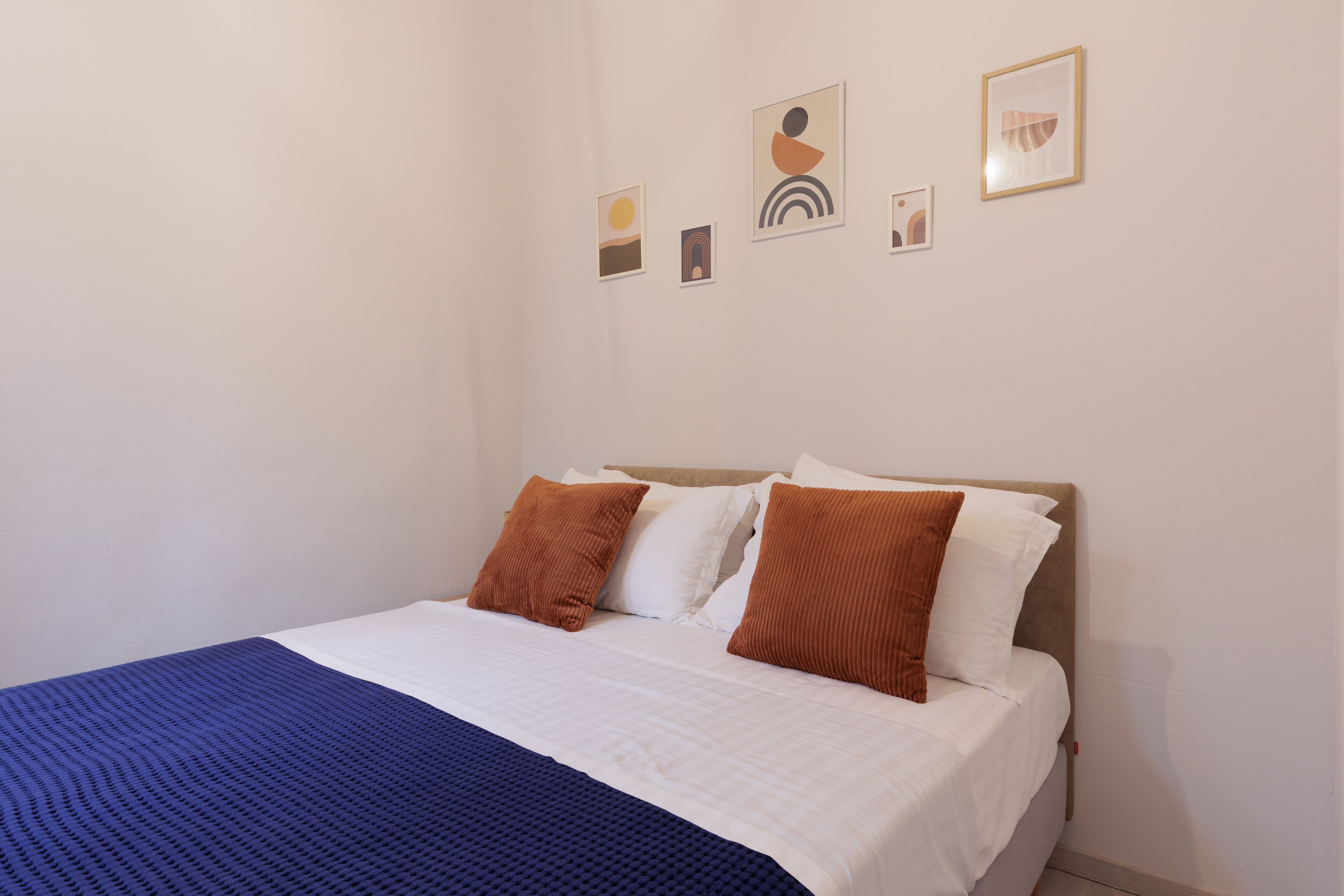 A neatly made bed with white and brown bedding, a blue blanket, and a headboard against a plain wall with four framed artworks.
