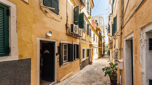 Ruada de paralelepípedos estreita, com edifícios tradicionais, uma combinação de venezianas abertas e fechadas, vasos de plantas e uma porta à vista.