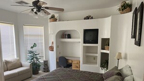 Une chambre à coucher comprenant un lit, un bureau, une chaise, un téléviseur et des étagères avec des plantes.