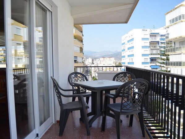 Un balcone con tavolo e sedie, affaccia su un panorama urbano con edifici e montagne.