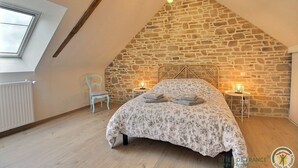 A bedroom with a sloped stone wall, a bed with floral bedding, two bedside lamps, a radiator, and a window with a skylight. - Leocadie - Plenee Jugon (PLENEE JUGON)