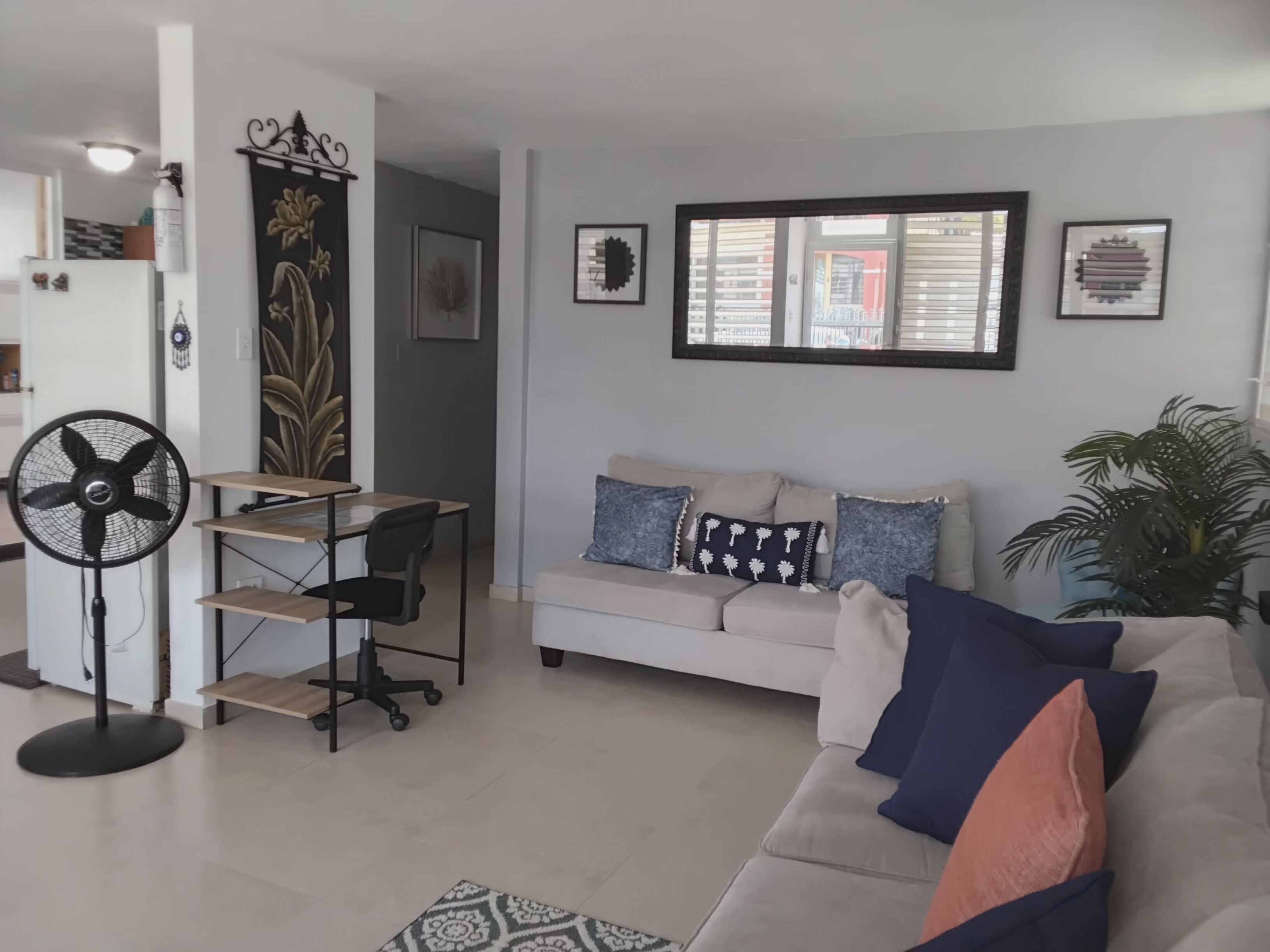 A living room with a white sofa, a standing fan, a desk with a chair, and a potted plant.