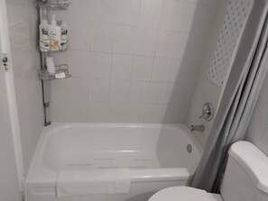 Un baño con una bañera blanca, un inodoro y una zona de ducha. Hay artículos de aseo en una estantería.