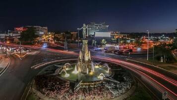 A nighttime cityscape with a central fountain, illuminated buildings, and a busy road with light trails.