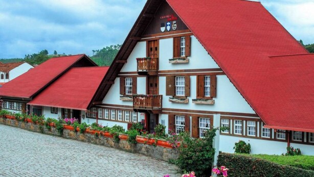 A traditional half-timbered building with a red roof and multiple windows, surrounded by cobblestone pavement and flower beds.