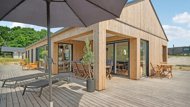 A modern wooden building with large glass doors and windows, a covered outdoor seating area with wooden furniture, and a wooden deck. - Stunning home in Samsø with sauna (Samsø)