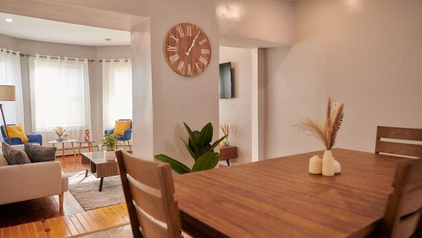 A modern living room with a wooden dining table, a large wall clock, and a sofa with yellow cushions. - Cozy apartment 1st 10min Walk Downtown | City View (Providence)