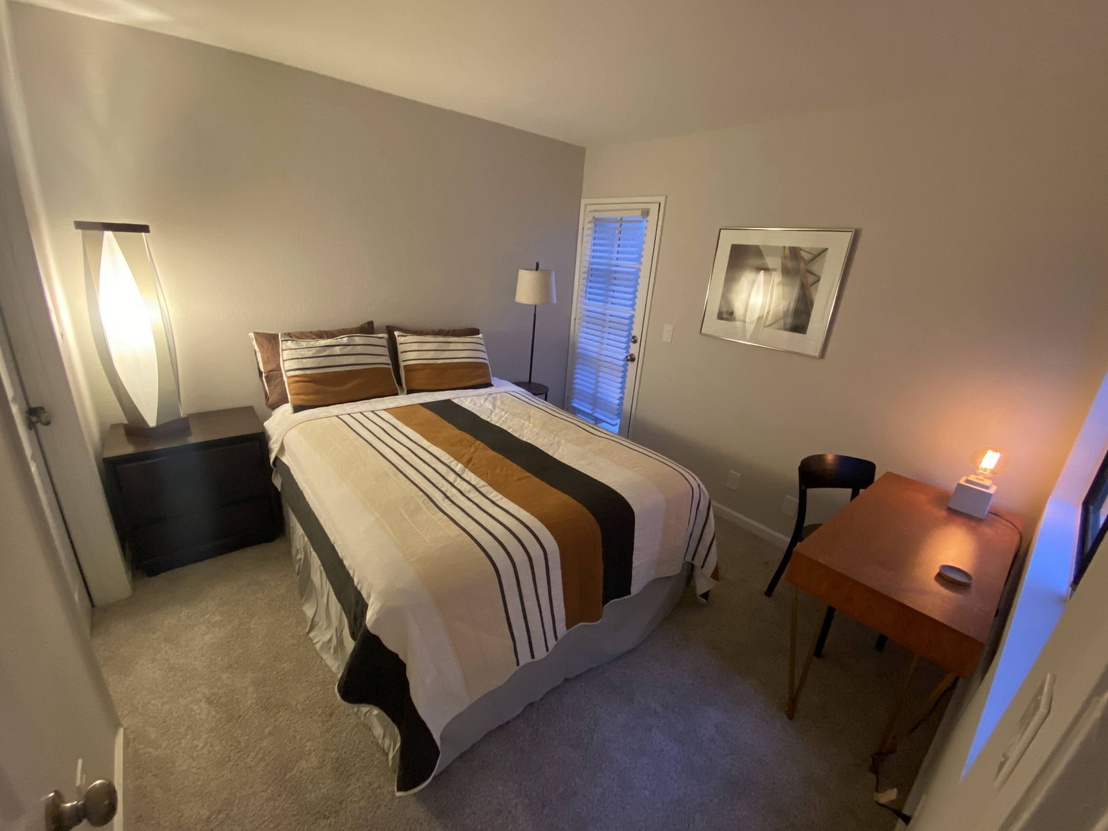 A bedroom with a bed, bedside tables, a desk, a chair, a lamp, a window with blinds, and a framed picture on the wall.