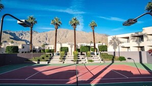 Un court de tennis avec une chaîne de montagnes en arrière-plan, des palmiers et des bâtiments modernes.