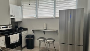 Une cuisine compacte avec des meubles blancs, un réfrigérateur en acier inoxydable, une machine à café et deux tabourets.