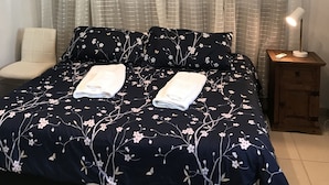 Un lit bien fait, avec une literie à motifs floraux, une lampe de chevet et une chaise dans une pièce éclairée par un plafonnier.
