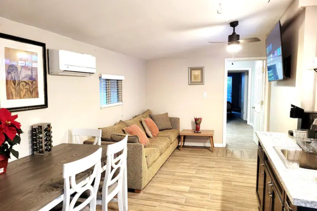 A modern living room with a wooden dining table, white chairs, a beige sofa with cushions, a small side table, and a TV mounted on the wall.