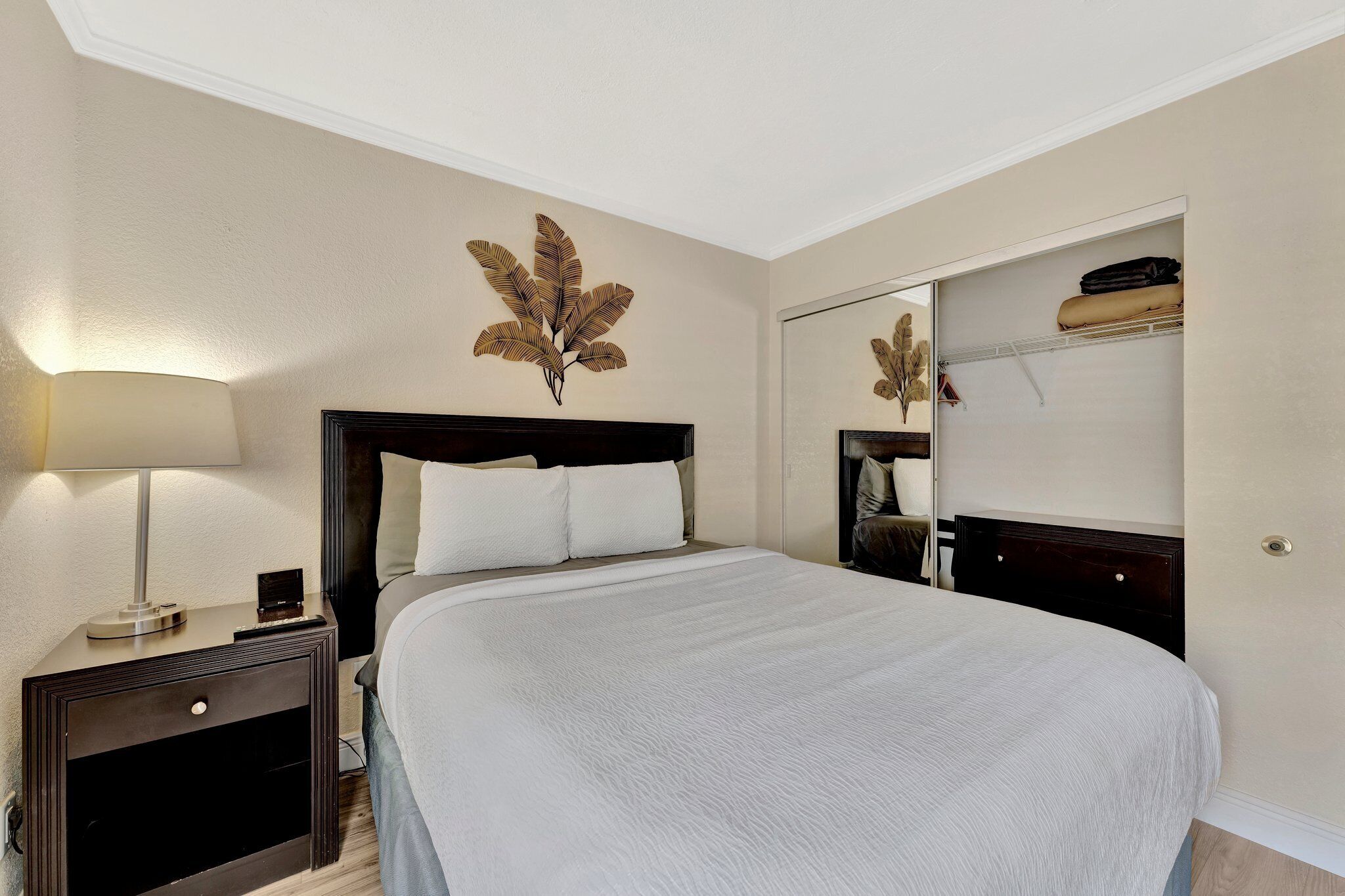 Una cama bien hecha con edredón blanco, una lámpara de noche y una hoja decorativa en la pared.