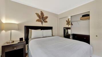 Una cama bien hecha con edredón blanco, una lámpara de noche y una hoja decorativa en la pared.