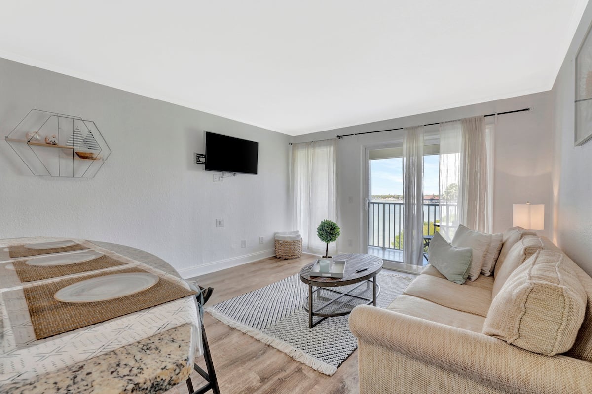 A modern living room with a sofa, a coffee table, a TV mounted on the wall, and a large window with curtains.
