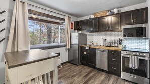 Una cocina compacta con armarios de madera oscura, un refrigerador de acero inoxidable, un horno empotrado y una ventana con cortinas.