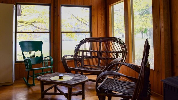 Una habitación con paneles de madera, una mesa, dos sillas, ventanas amplias con vista a árboles y un refrigerador.