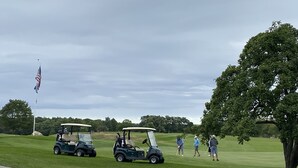 Un campo da golf con due golf cart, un alzata di bandiera con la bandiera americana e persone che giocano a golf.