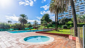 A swimming pool area with a circular hot tub, surrounded by a fence, palm trees, and a multi-story building in the background. - Gorgeous apartment in Estepona (Estepona)