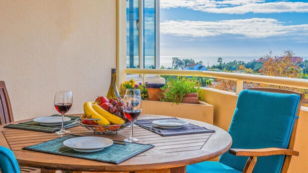 A round wooden table with a glass of red wine, a bowl of fruit, and a glass of white wine on a balcony overlooking a scenic view of the sea and buildings. - Gorgeous apartment in Estepona (Estepona)