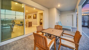Un espace patio avec une table et des chaises en bois, une porte vitrée donnant sur un espace intérieur et une vue sur l’extérieur.