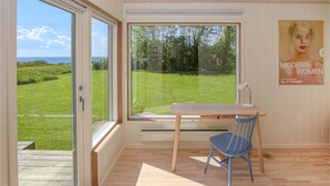 Et værelse med et stort vindue der giver udsigt over et grønt landskab og havet. Der er et træbord med en stol og en lampe.
