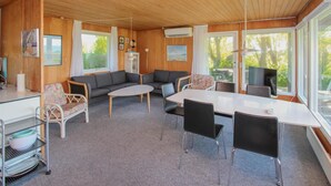 Indbydende hyttekabine med træloft, sofa, spisebord med stole, et køkken med køleskab og store vinduer med udsigt til naturen.