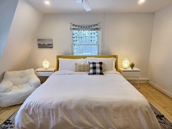 A bedroom with a large bed, a white chair, two bedside tables with lamps, a window with a view, and a small painting on the wall. - Red Bear Inn- 6.5 Baths! Grill, Pool, Central (Killington)