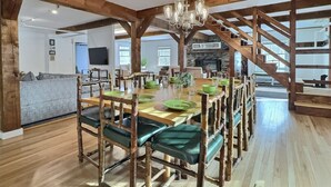 A spacious dining area with a wooden table set for six, surrounded by rustic wooden chairs with green cushions. A chandelier hangs from the ceiling, and a staircase is visible in the background. - Red Bear Inn- 6.5 Baths! Grill, Pool, Central (Killington)