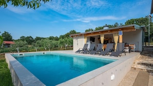 Una piscina rectangular con borde de azulejos, rodeada por una solana de hormigón. Seis sillones de descanso están alineados paralelos a la piscina, y hay una zona cubierta con techo de madera y una pequeña mesa. El entorno muestra un cielo azul despejado y vegetación al fondo.
