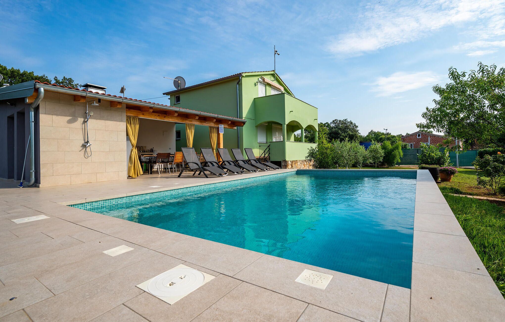 Ein Schwimmbecken mit gefliestem Sonnendeck, Liegestühlen und einer überdachten Sitzecke mit Tisch und Stühlen.