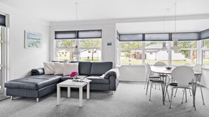 A modern living room with a grey sectional sofa, a white coffee table, and a dining area with white chairs and a table. Large windows with blinds provide views of the outside. - Lovely home in Samsø with WiFi (Samsø)