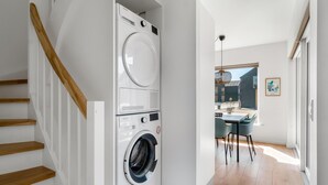 Een wasserette met gestapelde wasmachines en drogers, een houten trap en uitzicht op een eetgedeelte met tafel en stoelen.