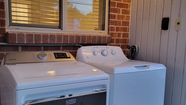 Deux machines à laver et deux sèche-linge Maytag blancs, placés côte à côte, avec une fenêtre au-dessus et un mur de briques sur le côté gauche.