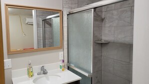 Une salle de bain avec un lavabo blanc, un miroir, une douche avec une porte en verre et des murs recouverts de carreaux gris.