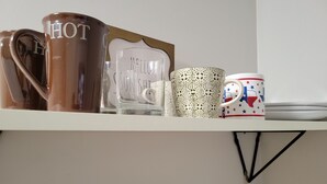 Une étagère avec divers mugs, dont un avec le mot « CHAUD » écrit dessus, un autre avec le mot « SOLEIL » et un mug en verre transparent.