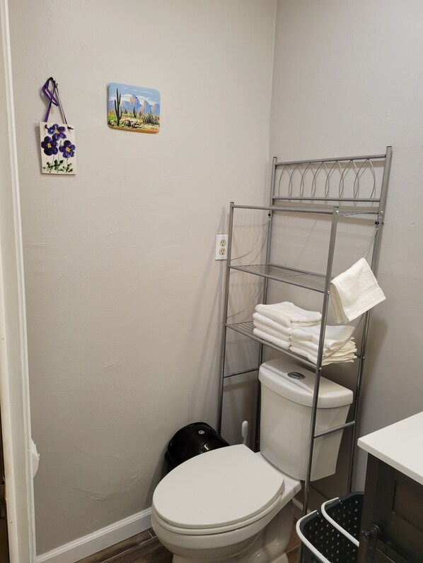 A bathroom with a white toilet, a metal shelf with towels, and two wall decorations. - Comfy Central Studio Guesthouse (Tucson)