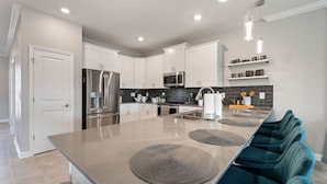 Une cuisine moderne avec des meubles blancs, des appareils en acier inoxydable et un grand îlot central agrémenté de chaises de couleur turquoise.