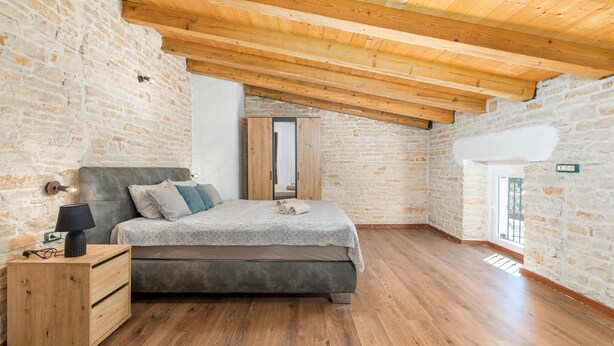 A bedroom with a wooden ceiling, stone walls, a bed with a grey headboard, a bedside table with a lamp, and a wooden dresser. - Awesome home in Modrusani (Modrusani)