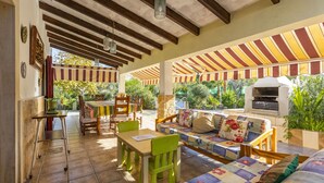 A covered patio area with a wooden ceiling, tiled floor, outdoor seating with a sofa and chairs, a table, and a grill. - Spacious 5-bedroom Villa with pool and barbecue (Can Picafort)