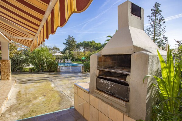 An outdoor patio with a stone floor, a barbecue grill, and a pool in the background.