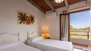 A bedroom with two beds, a nightstand with a lamp, a wall-mounted air conditioner, and a view of a balcony with a railing. - Spacious 5-bedroom Villa with pool and barbecue (Can Picafort)