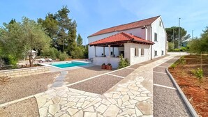 Une maison à deux étages, avec un toit recouvert de tuiles rouges, une piscine et un chemin de pierre menant à l’entrée.