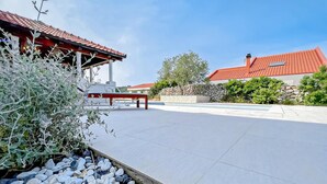 Un espace patio agrémenté d’un banc en pierre, d’une pergola et offrant une vue sur une maison dont le toit est recouvert de tuiles rouges.