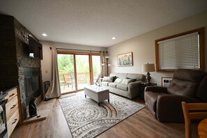 A living room with a fireplace, a sofa, a chair, and a large window with a view of the outdoors. - Schuss Village golf, pool, sleeps 6. 2 baths wood fireplace, outdoor pool summer (Mancelona)