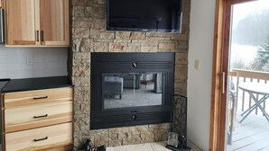 A kitchen with wooden cabinets, a stone wall, a black fireplace, and a view of a snowy outdoor deck.