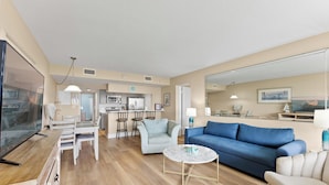 A modern living room with a blue sofa, a white coffee table, and a dining area with a kitchen in the background. - Direct Oceanfront Condo W/ Private Balcony, Heated Pool & Spa-like Hot Tub ☀️ (Cape Canaveral)