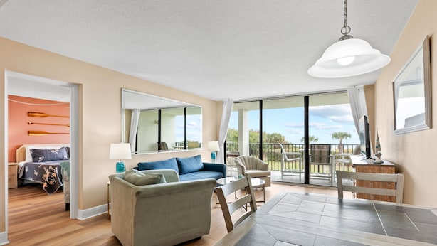 A spacious living area with a large sofa, a dining table, and a balcony with a view. - Direct Oceanfront Condo W/ Private Balcony, Heated Pool & Spa-like Hot Tub ☀️ (Cape Canaveral)