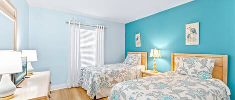 Un dormitorio con dos camas, una mesita de noche con lámpara, un espejo y una ventana con cortinas.